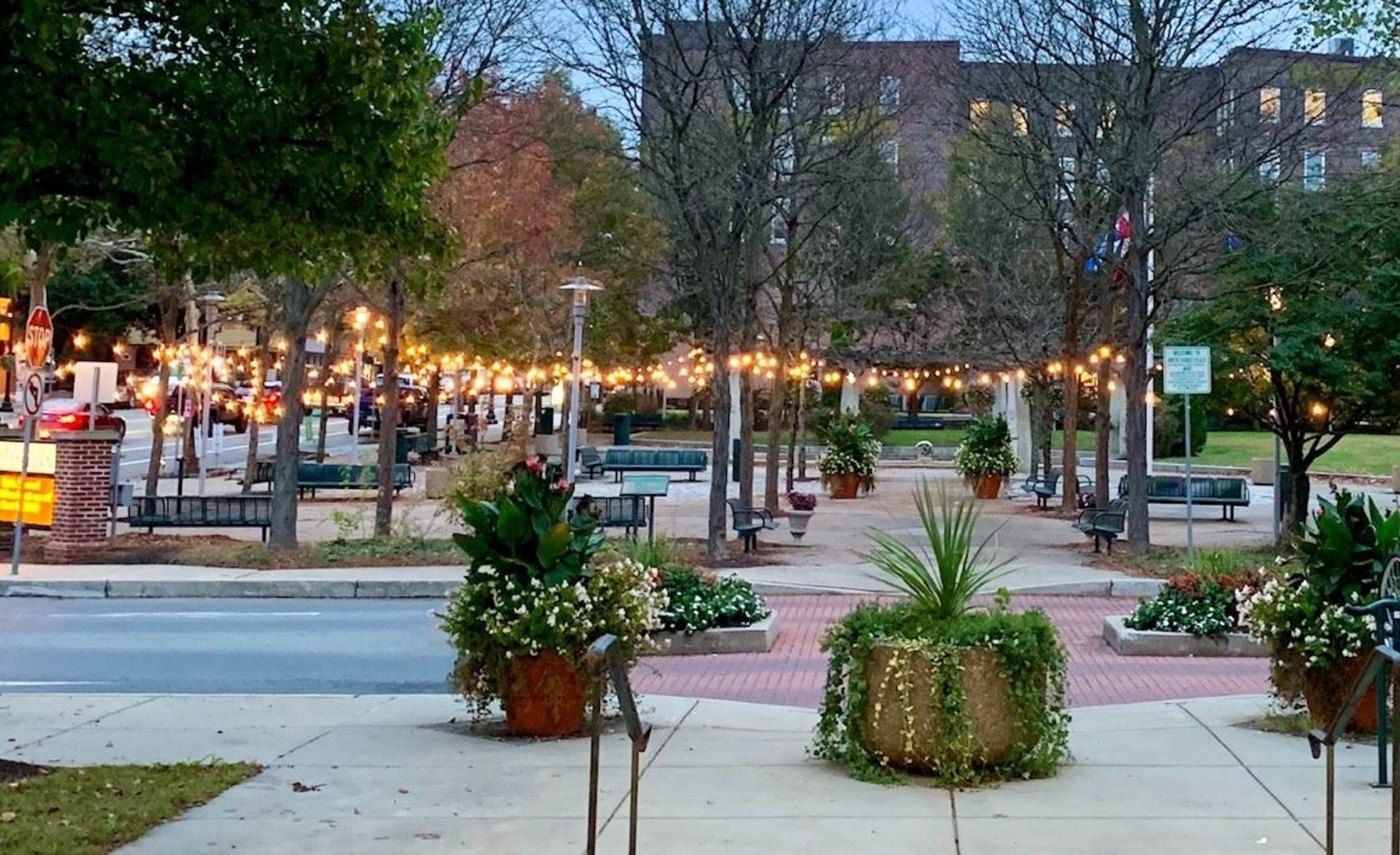String lights over the brick plaza of a small Pennsylvania downtown at dusk, with planters, benches, and storefronts.